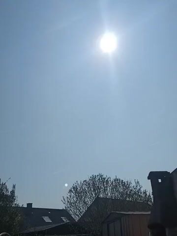 The video shows a bright, sunny day. The sun is high in the clear blue sky. Below the sky, there are rooftops of houses, trees, and a chimney stack. The scene is still and quiet. There are no people visible in the frame.
