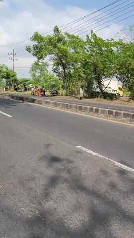 The video pans across a roadside scene in Siring, East Java. The time of day appears to be late morning or early afternoon, with bright sunlight casting shadows. The weather seems clear and sunny, with a blue sky dotted with a few wispy clouds.

The foreground shows a two-lane asphalt road with a white dividing line. The road appears to be in moderate condition, with some visible cracks. On the right side of the road, there is a concrete curb and a sidewalk area. Beyond the sidewalk, there is a line of trees with lush green foliage, providing shade. In the distance, there are more trees and some structures, possibly houses or small shops, and utility poles with overhead wires.

Several people are visible on the sidewalk and near the road. In the middle ground, a group of individuals are gathered around what appears to be a motorcycle and a cart. Some are standing, while others are near the vehicles. A person in a light-colored shirt and dark pants is walking along the sidewalk. Another person in a light shirt and possibly dark pants is seen further down the sidewalk. A motorcycle is parked on the side of the road.

The overall atmosphere is one of a typical day in a semi-rural or suburban area. There are no overt signs of distress or urgency. The activity suggests people going about their daily routines, possibly traveling or engaged in some form of commerce or social interaction. The presence of trees and the open sky contribute to a natural, outdoor setting. No text is visible in the media file.