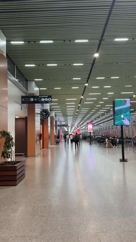 The image depicts a long, well-lit interior corridor, strongly resembling an airport terminal or a large public transportation hub. The perspective looks down the length of the hall, which features a light gray, speckled floor and a distinctive green-grey slatted ceiling fitted with numerous rectangular recessed lights and spotlights.

On the left side of the foreground, there is a large, square wooden planter holding a small green plant. Further along the left wall, a black directional sign with white arrows and pictograms (including a person with luggage, an airplane, and possibly restrooms) is visible, with some Arabic text. Beneath the sign, a round wall clock with a dark face and light hands is mounted. The wall also features wood-paneled sections and doorways.

The wide corridor is populated by several individuals and small groups of people. Some are walking towards the viewer, others are moving away, and a cluster of people is seated in a waiting area on the right side of the frame, further down the hall. On the right, large digital display screens on stands are visible, with one prominent screen showcasing a green landscape image. Additional red banners or signs are visible further into the distance.

The lighting is bright and artificial, casting soft shadows. The overall atmosphere appears calm and orderly, characteristic of a modern transit facility in Nouaceur, Morocco.