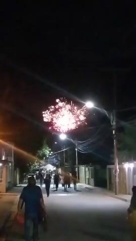 A nighttime street scene with fireworks exploding in the sky. People are visible walking on the street, some are gathered near tents in the background. There are also horses on the street. Streetlights illuminate the scene, and power lines are visible overhead. The atmosphere appears festive, with the fireworks suggesting a celebration.