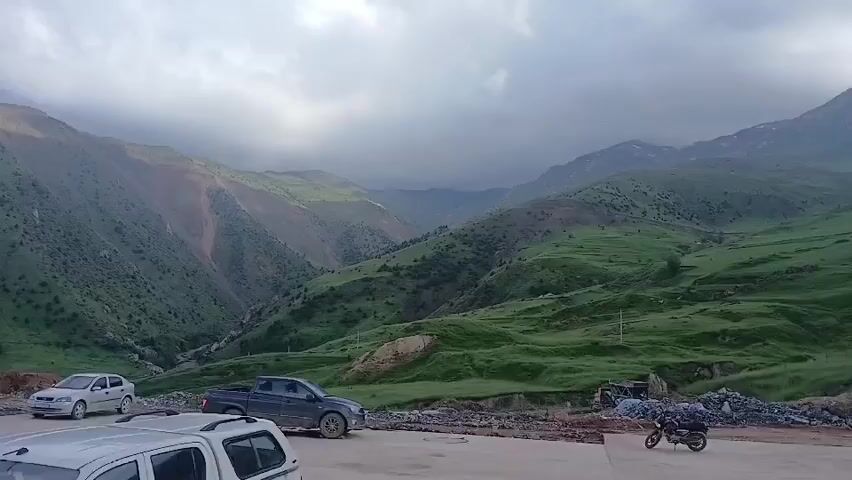 The video captures a panoramic view of a mountainous landscape under a cloudy sky. In the foreground, several vehicles are parked on a paved surface. These include a white hatchback, a dark grey pickup truck, and a white SUV in the foreground. Further to the right, a motorcycle is parked. Beyond the vehicles, the terrain slopes upwards into rolling green hills, with some areas appearing to be under construction or land excavation, evident from piles of dirt and debris. The hills are covered in lush green grass, with scattered patches of exposed earth and rock. In the distance, rugged mountains rise, with some peaks showing signs of snow. The sky is filled with dramatic, dark clouds, suggesting an overcast or possibly stormy weather condition. The overall scene conveys a sense of remote, rugged natural beauty, with a hint of human activity due to the presence of vehicles and construction-related materials.