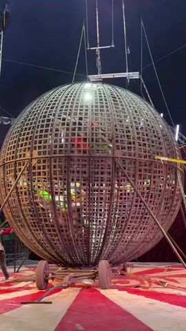 The video shows a large, spherical metal cage suspended in the air over a red and white striped circus-like floor. Inside the cage, several people wearing colored outfits are riding motorcycles. The motorcycles are moving around the inside of the sphere. The cage is supported by cables and metal poles. A light shines from the top of the cage. The background is dark.