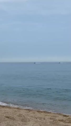 The video captures a serene beach scene with a pale blue sky and calm, blue water. The foreground is dominated by a sandy beach with small waves lapping at the shore. In the distance, two jet skis are visible on the water, creating small wakes as they move. The overall impression is one of tranquility and a peaceful day by the sea.