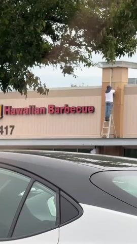 A person wearing a white shirt, blue jeans, and a dark hat stands on a ladder and appears to be painting a tan column on the exterior of a building. The building has a sign that says "Hawaiian Barbecue" in red letters. A car is parked in the foreground, obscuring the lower portion of the scene. The sky is overcast.