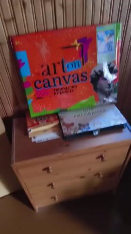 The video shows a person's hand with red nail polish reaching towards a large red art kit titled "art on canvas". The kit features a stylized number 7, the words "творчество на холсте" (creativity on canvas), and an image of a statue's head wearing pink glasses. To the right of the main kit is a smaller image of abstract art. Below the main kit, there are two smaller books or boxes, one with cartoon animals on it and the other with Russian text that appears to say "для рисования" (for drawing). The background consists of vertical wooden planks. The person's hand moves across the frame, briefly obscuring the art kit, and then points towards the smaller image of abstract art on the kit.