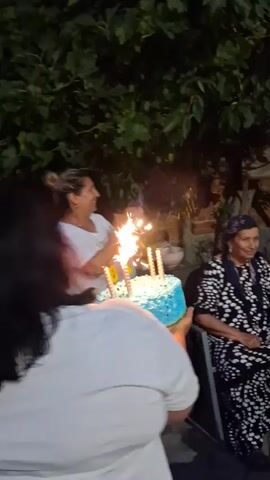 A birthday celebration is underway outdoors at dusk. In the foreground, a person in a white shirt holds a blue birthday cake with lit sparkler candles that spell out "84". An older woman wearing a headscarf and a black and white patterned dress is seated, smiling, and looking at the cake. To her right, a man with a shaved head and a beard, wearing a white t-shirt, stands and looks towards the cake. Another man, also with a shaved head, wearing a blue plaid shirt, is seated to the right. The background shows foliage and trees, suggesting a backyard or garden setting. The atmosphere appears festive and celebratory.