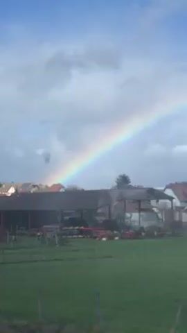 The video shows a rainbow stretching across the sky over a rural landscape. The rainbow's colors are distinct and visible against a backdrop of scattered clouds and blue sky. Below the rainbow is a scene with houses, buildings that look like barns, and open green fields. Some farm equipment, possibly tractors, are parked near one of the structures. The scene is shot from a slightly elevated position, perhaps from a moving vehicle or a building window. The foreground includes some tall grasses or reeds. The overall setting is peaceful and suggests a quiet, countryside environment.