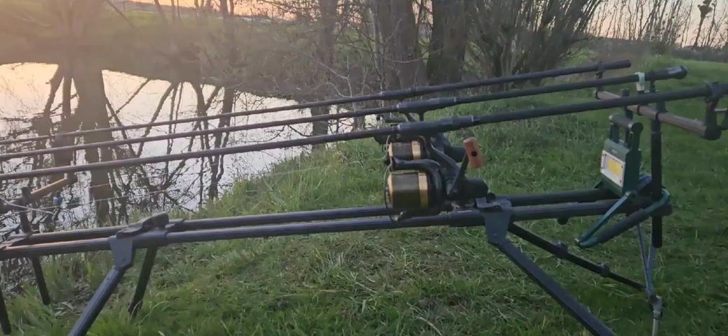 Here's a description of the video:

The video shows a fishing setup at a lake or river during sunset. The camera focuses on a black rod pod with three fishing rods and reels. The rods extend towards the calm water, reflecting the silhouettes of trees and the sunset sky. The background features trees and greenery along the bank. In the distance, there is a glimpse of a hill and what appears to be a building with smoke stacks.