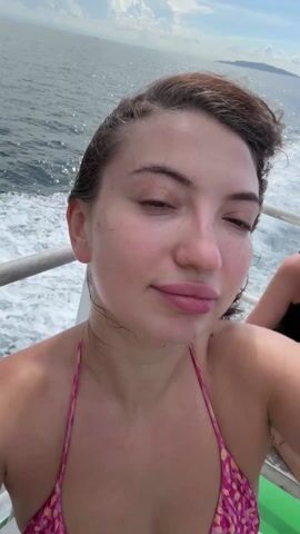 The video shows a woman on a boat, taking a selfie. She is wearing a pink and orange bikini top and white pants. The ocean is visible in the background, and there are other people on the boat. The woman puts her hair up and makes a kissing face. The boat is moving, creating white foam on the water.