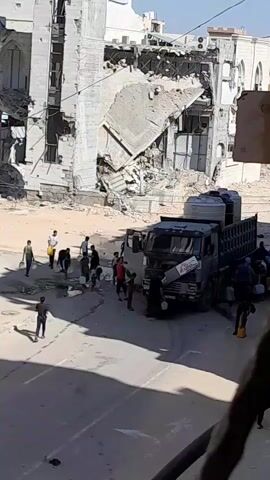 A group of people are gathered around a damaged building and a truck. Some individuals are carrying plastic containers, possibly for water. The building appears to have sustained significant damage, with debris scattered around. A truck, which is dark in color, is parked in the street. The sky is bright, suggesting it is daytime. The overall scene depicts a situation of distress or a humanitarian effort.