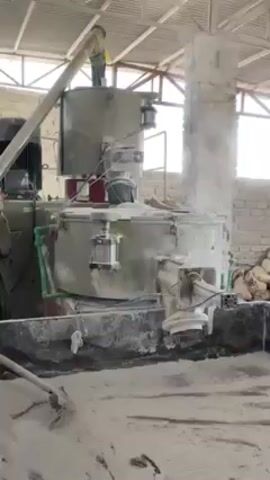 In an industrial setting, a large mixer is the focus, heavily coated with a light-colored powder. A person stands on top of the mixer, appearing to adjust or inspect it. Another person is seen shoveling the powder on the ground near the mixer. The environment is dusty and the light-colored powder is prevalent throughout the space. The mixer is connected to a large motor and various pipes. Bags of the same powder are stacked against a brick wall in the background. The roof structure is visible overhead, with a corrugated metal exterior.