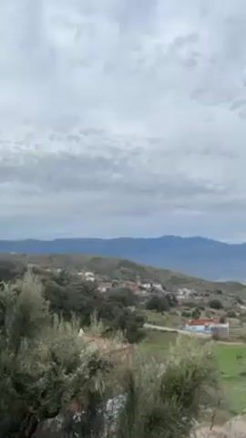The video shows a wide shot of a rural landscape under a cloudy sky. The foreground features green fields with scattered trees and a few small houses. In the midground, there are rolling hills with a cluster of buildings that seem to be a small village. The background shows a range of distant mountains fading into the horizon. The camera slowly pans from left to right, revealing more of the landscape.