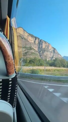 The video shows a view from inside a moving vehicle, likely a bus, looking out the window. The foreground on the left shows part of a seat with patterned upholstery and a headrest. The window reflects the interior of the vehicle and the passing scenery. The middle ground features a road curving to the right, bordered by a metal guardrail and vegetation. A lamppost is visible along the roadside. In the background, a large, steep mountain with rocky, textured slopes dominates the landscape under a clear blue sky. The sun appears to be low, casting a warm light on the mountain. The overall impression is a scenic journey through a mountainous region.