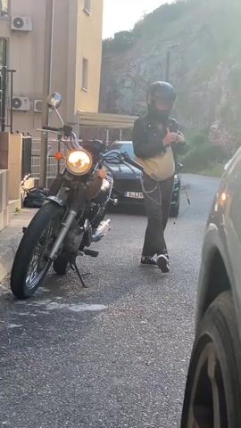 A person wearing a black helmet, a black leather jacket, dark patterned pants with a silver chain accessory, and black and white sneakers stands on a paved surface. They are adjusting their jacket and a beige fanny pack while standing next to a parked motorcycle. The motorcycle, possibly a retro style, is on its kickstand, and its headlight is illuminated, suggesting the engine is running. In the background, a black Audi car with a Slovenian license plate "S LJ910" is visible. Further back, there are buildings with air conditioning units and a rocky, mountainous landscape under bright, possibly late afternoon, sunlight. A part of another vehicle is visible in the immediate foreground on the right. The scene appears to be an outdoor, narrow street or driveway in a residential or semi-rural area.