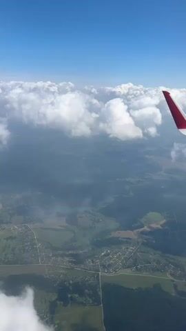 The video shows a view from an airplane window looking down at the earth. The sky is blue with fluffy white clouds. Below the clouds, there is a landscape of green fields, forests, and scattered houses. Part of the airplane wing, which is red and white, is visible in the upper right corner of the frame. The camera is moving slightly, suggesting the plane is in motion.