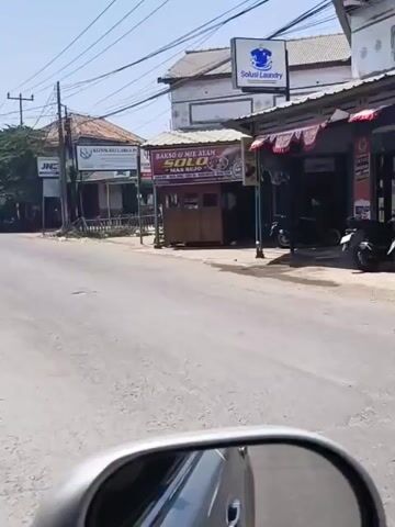 The video shows a street scene with several buildings and vehicles. In the foreground, a side mirror of a vehicle reflects the road ahead. A road stretches into the distance, with a few cars and motorcycles visible. On the right side of the road, there are several shops, including a laundry service, a food stall selling "Bakso & Mie Ayam Solo," and what appears to be a convenience store. Two people in school uniforms are riding a motorcycle, with one driving and the other sitting behind. On the left side of the road, there is a clinic and another shop. The sky is clear and bright, suggesting a sunny day. The overall scene depicts a typical street in what appears to be a commercial area, likely in Indonesia, given the signage and the presence of motorcycles.