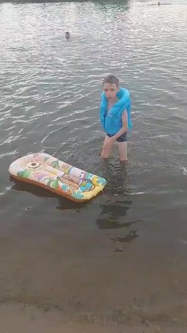 A young boy wearing a blue life vest and dark shorts wades in shallow, murky water near the shore. He is reaching towards an inflatable raft decorated with cartoon characters, which is floating on the water in front of him. The raft is yellow and white with orange trim and features images of people. In the background, other people are visible in the water, including two children and an adult in the distance, seemingly playing or swimming. The environment appears to be a natural body of water, possibly a lake or river, with a sandy or muddy shoreline. The lighting suggests it is daytime, possibly overcast.