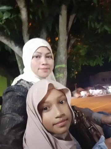 A young woman wearing a light pink hijab and jeans is sitting outdoors at night. She is looking towards the camera and smiling. In the background, there is a gas station with several motorcycles lined up. The scene is illuminated by artificial lights.