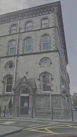 This media file captures a street scene in Waterford, Ireland, on a cloudy day. The perspective is from a moving vehicle, looking towards a tall, old stone building with arched windows. In front of the building, a black metal fence lines the sidewalk.

Further down the street, a lamppost displays a blue banner with a white "P" symbol, indicating parking, and the word "WATERFORD" written in white text above it. The street is lined with trees and more buildings, and there is traffic with several cars visible. In the distance, a large, grey metal structure, possibly a bridge or crane, can be seen against the sky.

The overall mood is calm and ordinary, depicting a typical day in the city. The sky is overcast with some scattered clouds, suggesting it might be daytime but not particularly sunny. There are no prominent people or specific events captured, just the general atmosphere of a city street.