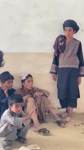 The video shows a group of five young boys in what appears to be a classroom or similar indoor space. The walls are bare and somewhat dirty, and the floor is dirt or concrete. The boys are dressed in traditional clothing. One boy stands slightly apart from the others, wearing a black tunic with intricate embroidery and a black turban. He has a brown cloth draped over his shoulder. The other four boys are seated on the floor, leaning against the wall. One boy in the foreground is wearing a light-colored tunic and is looking directly at the camera with a pensive expression. To his left, another boy in a tan tunic with a white cap is looking towards the standing boy. Further back and to the left, two boys in darker tunics are also seated, one with a black cap. The lighting suggests it is daytime, possibly with natural light coming from an unseen source. The overall impression is one of a candid moment captured, with the boys appearing to be waiting or observing something.