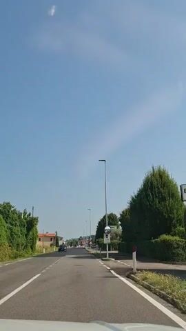 The video shows a view from a vehicle driving down a paved road. The road has white dashed lines marking the lanes. On the left side of the road, there are houses and greenery. On the right side of the road, there are more houses, trees, and a street lamp with a speed camera mounted on it. The sky is clear and blue with a few faint clouds. The sun is shining, creating shadows on the road. A black car is visible in the distance on the left side of the road. The vehicle appears to be moving at a moderate speed.