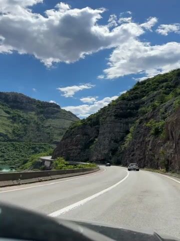 The video shows a road winding through a mountainous landscape on a bright, sunny day. The road is paved and has a solid white line down the middle. On either side of the road are steep, rocky hills covered with green vegetation. In the distance, more mountains can be seen under a blue sky with fluffy white clouds. Two cars are visible on the road, one in front and one further ahead. The video is shot from the perspective of a vehicle traveling on the road, with a portion of the car's dashboard visible at the bottom of the frame. The sound of wind is audible in the video.