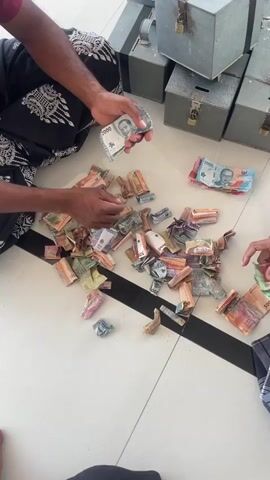 The video shows an overhead view of several people counting money on a tiled floor. The money is a mix of different denominations and colors, scattered in a pile. The people are dressed in traditional clothing, and they are using their hands to sort and count the bills. In the background, there are several metal boxes with locks, which appear to be donation boxes. The lighting is bright, suggesting an indoor setting.