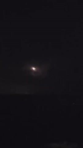 A time lapse video shows a dark, cloudy sky over a body of water. Lightning flashes in the distance, illuminating the clouds. The scene appears to be a storm over the ocean at night.