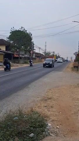 A black car is driving down a two-lane road. There are other cars and motorcycles on the road. There are trees and houses on the side of the road. The sky is cloudy.