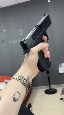 The media file shows a close-up of a person's hand holding a black handgun. The hand is wearing a silver and gold watch and a large silver ring. The person has tattoos on their arm, including one that appears to be a cartoon bell. The background is a dark gray wall with some faint white lettering that says "rez:monta" and an apple logo. There is also a red object, possibly furniture, visible to the left, and a black microphone stand to the right. The flooring appears to be tiled. The lighting suggests an indoor setting, possibly a workshop or studio. The overall mood is unclear due to the focus on the object. The time of day and weather are not discernible. The visible text "rez:monta" might indicate a business name or a slogan.