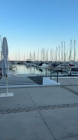 The video shows a marina with many sailboats docked under a clear blue sky. In the foreground, a zebra crossing is visible on a paved walkway. A large white umbrella is positioned to the left of the frame. A young person on a red bicycle with a black helmet rides past the zebra crossing. Further in the background, a person is seen walking near the water. The overall environment is a calm and sunny day at the harbor.