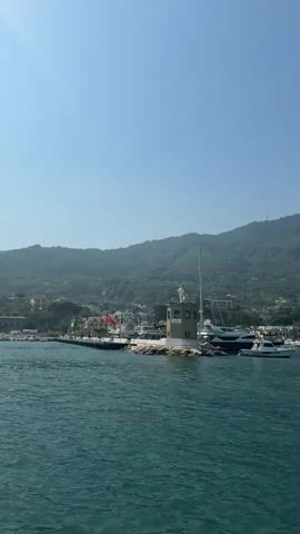 The video pans across a coastal town with a lighthouse and several boats docked. The water is calm and blue, reflecting the clear sky. The town is built on a hillside covered in lush green vegetation. The lighthouse is small and cylindrical with a white top and a statue of a figure on it. There are several yachts and smaller boats moored in the harbor, as well as some buildings visible on the shore. The camera pans from left to right, revealing more of the coastline and the mountains in the background.