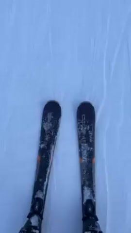 The video shows a person skiing down a snowy slope. Initially, the skis are visible against the white snow, then the camera shows the person's face, sticking out their tongue and wearing a helmet and ski goggles with a red hooded jacket. The camera shifts to show the snowy slope ahead, bordered by trees and a mountainous backdrop in the distance. The scene is bright, indicating a clear day.