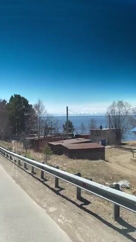 The video shows a view from a moving vehicle driving along a road with a metal guardrail. Beyond the guardrail, there is a strip of dry grass and several small, dilapidated buildings with rusty roofs. Further in the distance, a body of water is visible, with faint, snow-capped mountains visible on the horizon. The sky is a clear, bright blue. There are trees, some deciduous and bare, and some evergreen. The general impression is of a slightly neglected, rural or industrial landscape on a clear, sunny day.