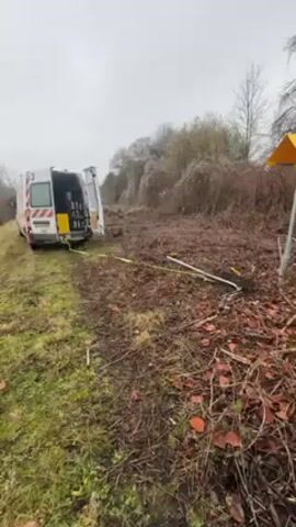 The video shows a white van with its rear doors open, parked on a grassy area next to a dirt field overgrown with bushes and dead vegetation. A thick, yellow cable extends from the back of the van into the dirt field. Several white tubes are visible in the dirt. A yellow warning sign on a pole is to the right of the frame. The sky is overcast and the environment suggests a rural or roadside location. The purpose of the van and cable setup is not clear from the visual information alone.