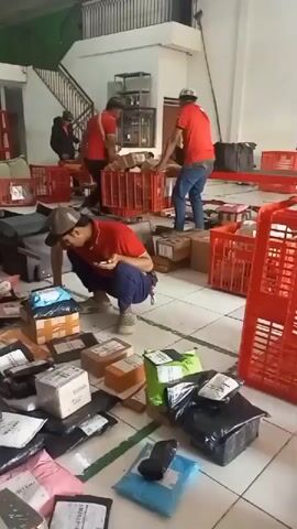 In an indoor warehouse or distribution center, several individuals in red shirts are seen handling numerous packages and red plastic crates. The floor is covered with many packages of various sizes and colors, some appearing to be in bags and others in boxes. Some individuals are sorting through the packages, while others are moving the crates. One person is crouched down, looking at a package. The environment appears busy and somewhat chaotic due to the large volume of packages scattered on the floor. There is a staircase visible in the background, and the walls are white.