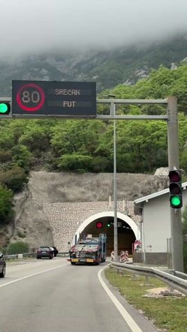 A car approaches the entrance of a tunnel. The tunnel is surrounded by green trees, mountains and low-lying fog.  A flatbed truck with a load is entering the tunnel.  Traffic lights, a speed limit sign of 80, an illuminated green arrow and a red X hang over the entrance.  A white building is next to the tunnel entrance. A black car is parked to the left of the tunnel.  The camera approaches the tunnel.