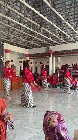 Four young women are performing a traditional dance indoors. They are wearing matching red tops with ornate collars and cuffs, and striped sarongs. Each dancer has a red scarf draped over their shoulders and is holding a red fan in each hand, which they are using as props in their choreography. The dancers are spaced apart on a tiled floor. In the background, there is an audience seated on chairs, also mostly wearing red. The room has a high, exposed metal ceiling and red-and-white striped pillars. There is a large black speaker on the left side of the frame. The overall atmosphere appears to be a cultural event or performance.