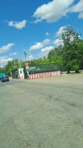The video shows a view from inside a vehicle, looking out at a road under a bright blue sky with scattered clouds. In the foreground, the dashboard of the car is visible. The road appears to be a two-lane asphalt road passing through a semi-rural area with trees and greenery on either side.

At the beginning of the video, there are some structures on the right side of the road, possibly a checkpoint or a construction area, with barriers and signage. A white van is visible in the distance. As the video progresses, the perspective shifts, showing more of the road and the surrounding trees.

Later in the video, the car appears to be approaching a curve in the road. The environment remains green and wooded. Several vehicles, including a white van and a blue car, are seen on the road. The presence of guardrails along the side of the road is also noticeable. The overall scene suggests a daytime drive through a well-maintained road in a natural setting.