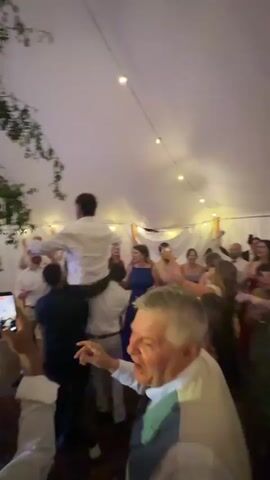 A group of people are dancing and celebrating inside a tent at what appears to be a wedding reception. One man is being lifted onto the shoulders of two other men and is dancing with his arms outstretched. String lights hang from the tent ceiling, and there are green branches hanging from the left side of the frame. Another woman in a blue dress is dancing in the background, and a man in a white shirt is seen holding a phone, seemingly recording the event. In the foreground, a man with gray hair is partially visible, facing away from the camera, with green lighting on his face. The overall atmosphere is festive and energetic.