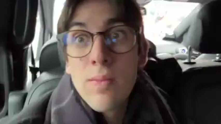 The video shows two people riding in the back seat of a vehicle. The camera is pointed at a young man with brown hair and glasses, who is sitting in the back seat. He is wearing a grey scarf. In the back seat next to him sits a woman with short brown hair and glasses. The car is driving on a highway, and the scenery outside the window is blurred. The young man looks at the camera.