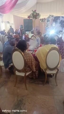 The video shows a large gathering of people sitting at tables in what appears to be a banquet hall or wedding reception venue. The room is decorated with white and pink draping and floral centerpieces.  Most people are seated, with some dressed in traditional Nigerian attire.  There are some people standing and moving around.  The camera is positioned among the guests, showing a partial view of the tables and the people seated at them. The floor is shiny and tiled.  Ceiling fans can be seen turning above.
