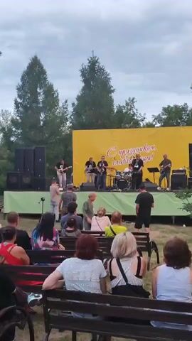 A band is performing live on an outdoor stage. The band consists of several musicians, including guitarists, a saxophonist, and a drummer. They are playing on a stage with a large yellow backdrop that has text written on it. In front of the stage, there is an audience seated on benches, watching the performance. The scene appears to be set in a park or an open area with trees in the background. The sky is cloudy, suggesting it might be overcast or late in the day. The overall mood seems relaxed and festive, typical of an outdoor concert or local celebration. The visible text on the backdrop is in Russian and translates to "Happy Holiday, Slavgorod!".