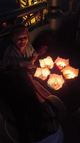 An elderly Asian woman sits in a dark environment, surrounded by several small, paper lanterns with lit candles inside. She wears a dark hat, a light blue shirt, and a dark patterned skirt. She is holding what appears to be money and smiles at unseen people. The bottom of a person’s leg is visible in the foreground, wearing light-colored pants. The scene appears to take place at night, possibly on a boat or near a waterway. The only illumination comes from the lanterns. The video includes voices expressing thanks.
