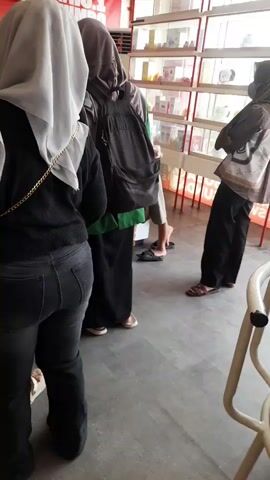 The video shows a line of people in what appears to be a store or food establishment. In the foreground, a woman wearing a black top, black jeans, and a white hijab is standing, with her back to the camera. To her left, a child's leg is visible. Behind her, another person wearing a grey hijab and a backpack stands. Further back, other people are visible, including someone at a counter with a computer screen. The environment features a red wall with signage and shelves stocked with products. The floor is a gray, tiled surface. The overall scene depicts a mundane moment of people waiting in a retail or service setting.