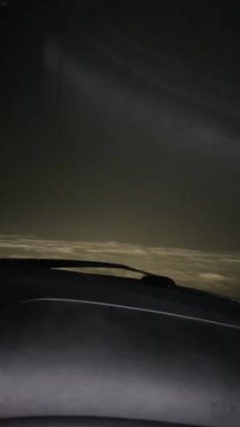 The video shows a car driving at night in heavy snow. The view is from the driver's perspective, looking out through the windshield. Snow is falling heavily, and visibility is poor. The wipers are on, but the snow is still accumulating on the windshield. The interior of the car is visible, including the dashboard, steering wheel, and rearview mirror. A white air freshener hangs from the rearview mirror. The dashboard lights are illuminated.