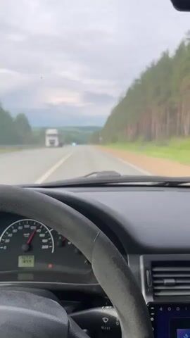 The video captures a first-person perspective from inside a car driving on a highway during the daytime. The sky is overcast with clouds, but the scene is well-lit.

The interior of the car is visible, including the steering wheel, dashboard, and a car infotainment system. The speedometer indicates that the car's speed varies, starting around 80 km/h and increasing past 100 km/h at different points. The odometer displays "155934" and "18.4". The infotainment system shows the word "Bluetooth" and appears to be playing media. A stalk behind the steering wheel has "RESET" printed on it.

Outside the car, an asphalt highway stretches into the distance. Tall pine trees form a dense forest lining the right side of the road. In the initial seconds, a large white truck is seen ahead on the road. Later, a black car passes in the opposite direction. A triangular road sign with a red border is briefly visible on the right side of the highway, near the forest. The landscape in the distance features gentle hills or plains under the cloudy sky. Music is playing in the background throughout the video.