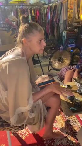 A video shows three people seated in a cluttered room, engaged in a conversation. The woman on the left, with her blonde hair tied up, is wearing a beige robe and is seated on a stool covered in leopard print fabric. Her legs are bare. Across from her is a woman with long, dark hair, wearing a gray t-shirt. To the right, a man with long blonde hair and a beard is dressed in a colorful floral robe. He’s wearing sunglasses. They are seated around a small wooden table laden with assorted items. In the background, a rack of clothing is visible along with fans and various decorations. The room is dimly lit, creating a cozy atmosphere. The floor is covered with a patterned rug. The camera briefly shifts its focus to show the interior of the room, emphasizing the cluttered and bohemian aesthetic.
