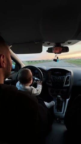 Inside a car, a baby sits on an adult's lap in the driver's seat, holding the steering wheel of what appears to be a Ford Focus. The adult, presumably the father, has his arm around the baby, and his back and head are visible from behind. The baby is wearing a light-colored long-sleeved top and white socks. Through the windshield, a road stretches ahead into the distance, flanked by fields, under a sky painted with soft orange and blue hues, suggesting late afternoon or early evening.

In the rearview mirror, another adult's face and hand holding a smartphone are reflected, likely the person recording the video from the passenger seat. A dark car air freshener hangs from the rearview mirror. A smartphone is mounted vertically on the dashboard.

An adult male voice can be heard speaking to the baby in Russian, saying, "You'll cut your hands, Daddy will cry," and then "What, Eva, you'll cut your hands..." The car appears to be moving slowly or is stationary, as indicated by the low speed on the dashboard. The scene captures a playful moment between a parent and a child.