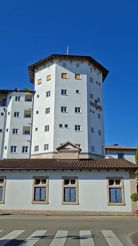 The video shows a white, multi-story building with a distinctive octagonal tower in the center. The building has many windows and a dark-tiled roof. A sculpture or decoration is visible on top of the octagonal tower. To the left of the tower, there is a taller, white building with balconies and numerous windows, connected to the main structure. In front of the building, there is a lower, white structure with arched windows. A hedge runs along the front of the property, and a street with a pedestrian crossing is visible in the foreground. The sky is clear and blue. The video pans slightly to the right. A stork's nest is visible on top of the tower.